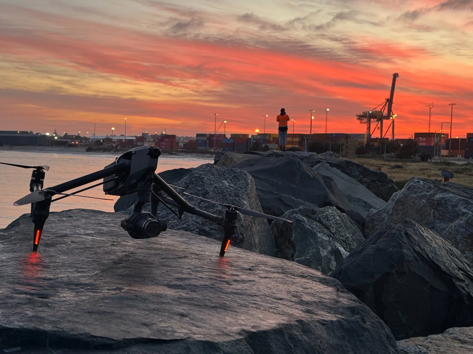 DJI Inspire 3 on a rock breakwater at sunrise while a crew member in hi-vis watches the sky, with a gantry crane and container stacks against a red dawn sky at Fremantle Port, Western Australia