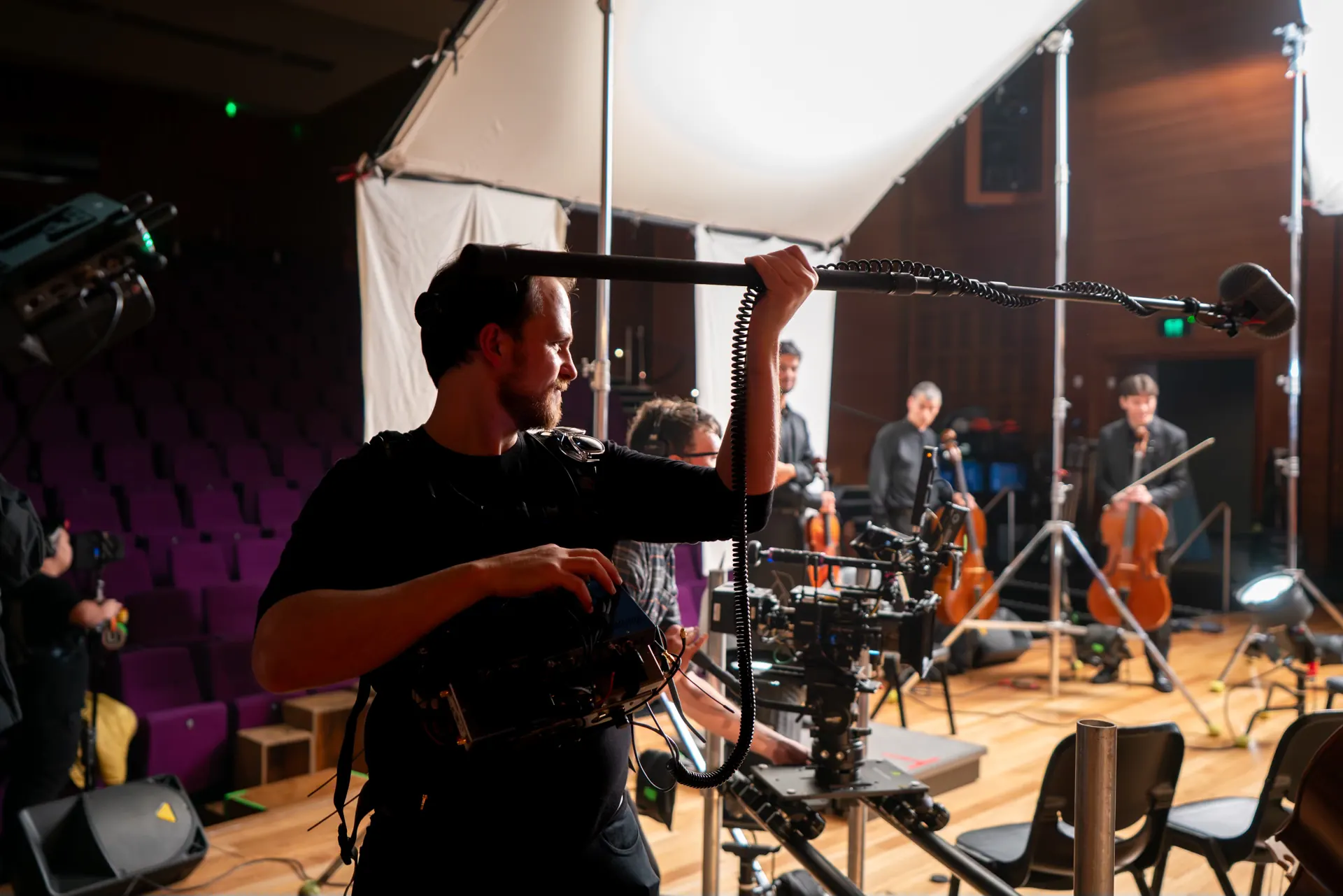 Richard Hubbard booming a string quartet on a concert-hall stage, bag-mounted mixer on his chest, with a camera on a slider in the foreground