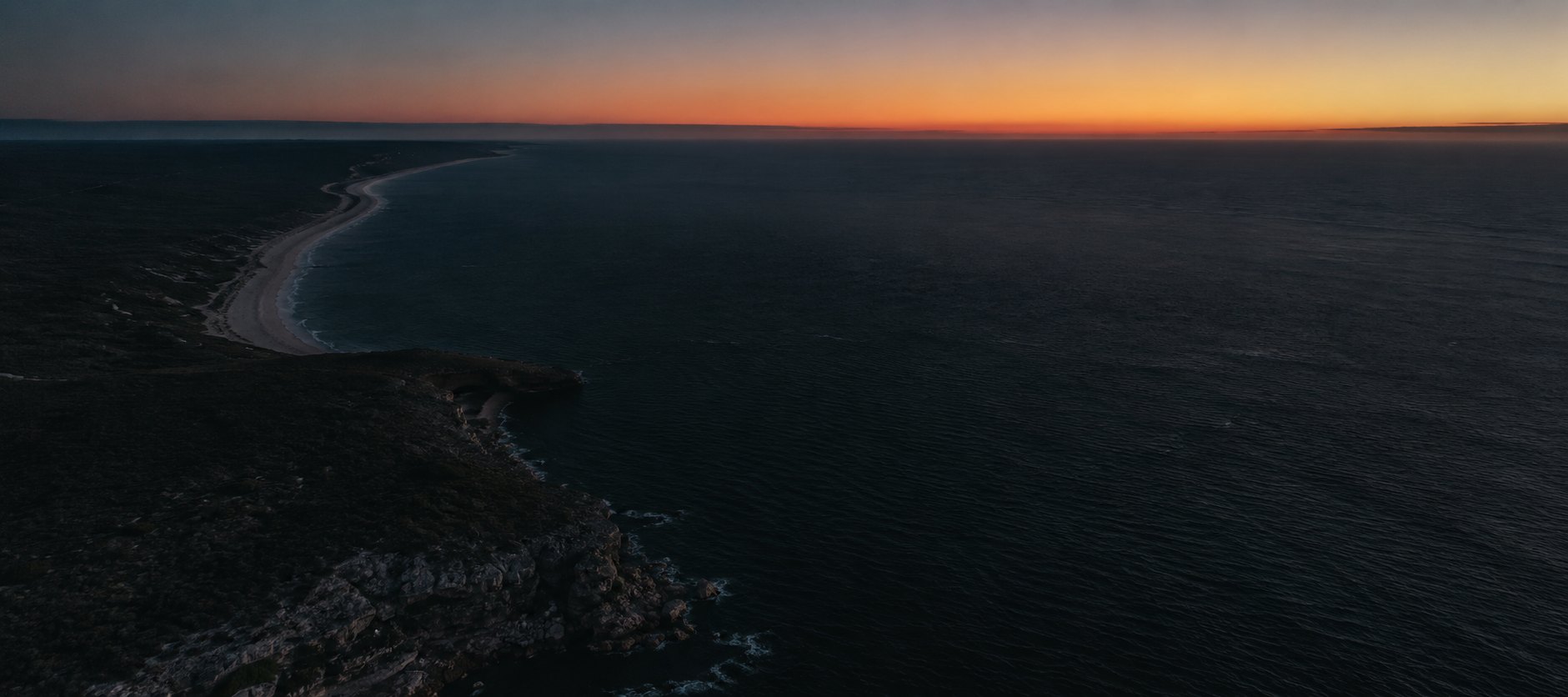 Aerial view along a remote Western Australian coastline at dusk, dark limestone headland and a long pale beach curving away beside the Indian Ocean