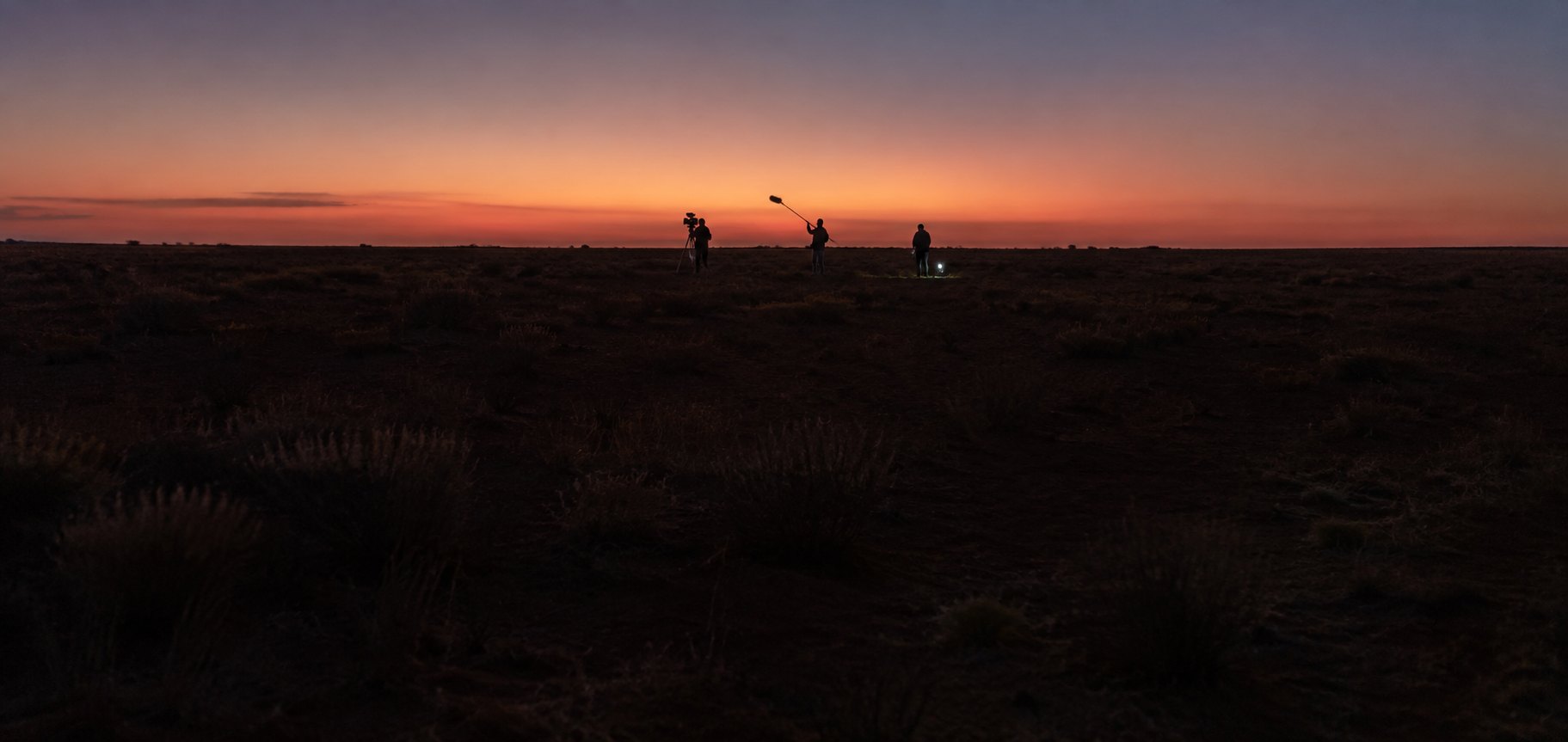 A small film crew silhouetted against a dusk sky on a flat outback plain, one operating a camera on sticks and one holding a boom pole raised high
